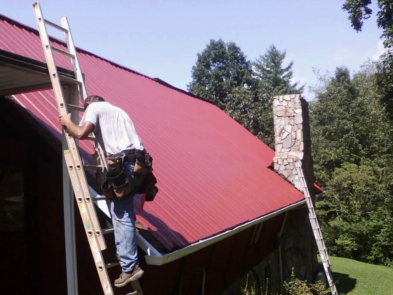 Roofer on a ladder beside a red metal roof and stone chimney — Soffit & Fascia Repair by Roofing Brothers in Tuttle